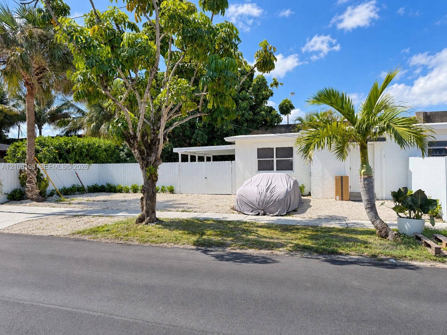 600 NW 116th Terrace, Miami, FL 33168 White exterior facade featuring a covered carport, gravel front yard, white perimeter fencing, and mature landscaping