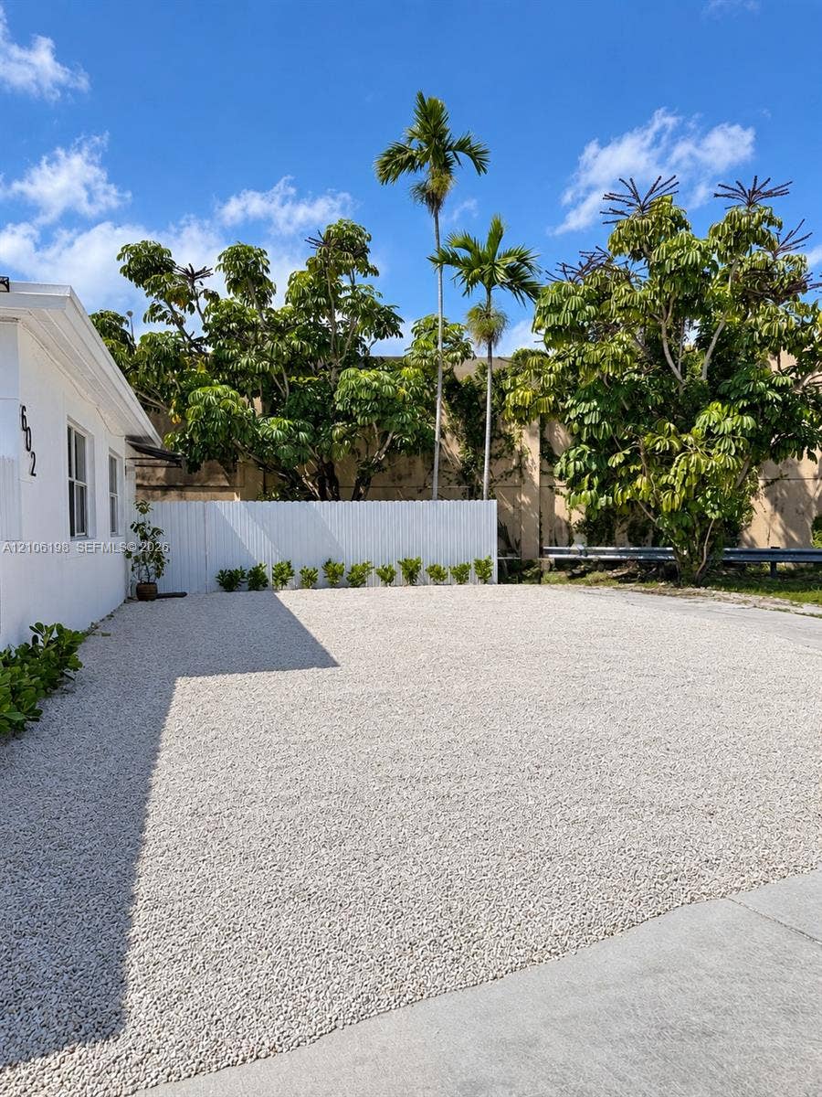 600 NW 116th Terrace, Miami, FL 33168 Expansive gravel parking area featuring a white privacy fence, low-maintenance landscaping, and mature palm trees