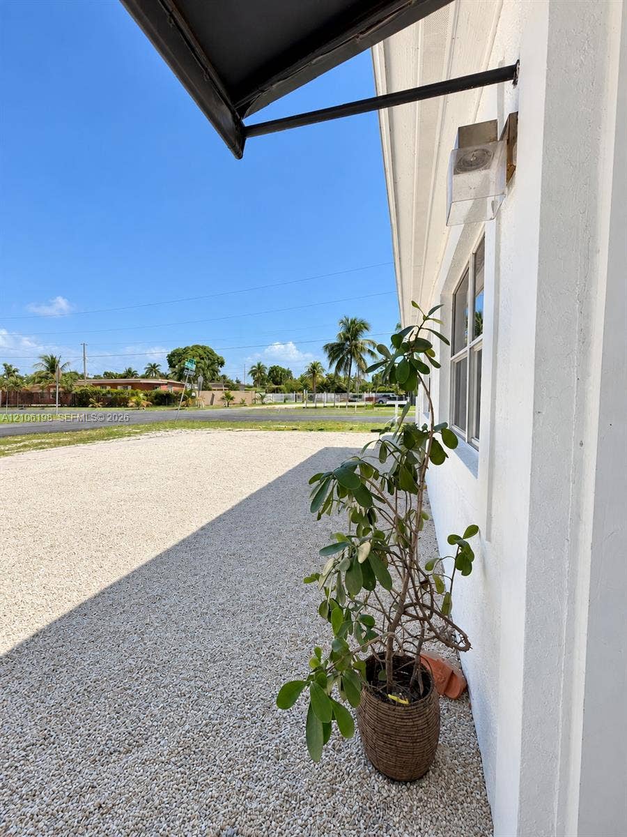 600 NW 116th Terrace, Miami, FL 33168 Exterior wall featuring white siding, a fixed-pane window with a frame, an attached metal awning, and an exterior wall-mounted light fixture, complemented by a gravel surface