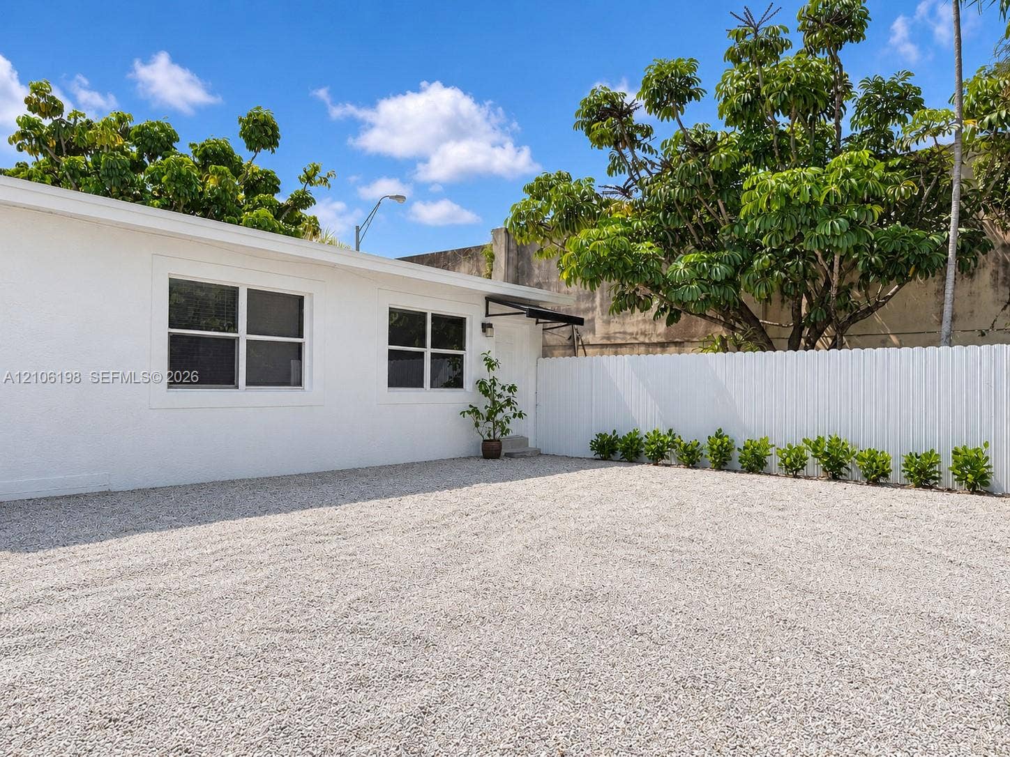600 NW 116th Terrace, Miami, FL 33168 Exterior featuring a white stucco facade, gravel ground cover, and a white privacy fence accented by mature landscaping