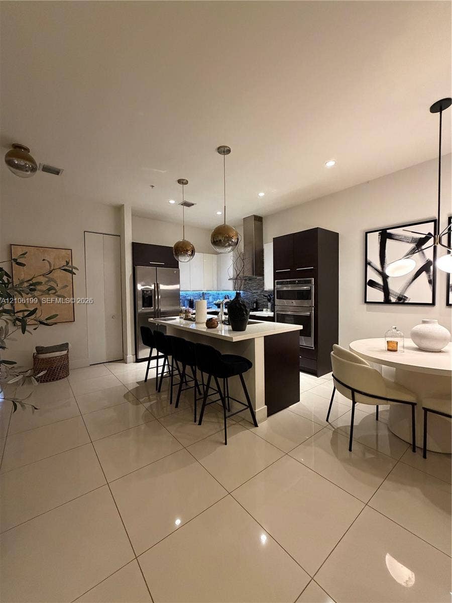 Modern kitchen featuring flat-panel cabinetry, a central island with stone-surface countertop, stainless steel double wall oven, stainless steel refrigerator, and large-format tile flooring
