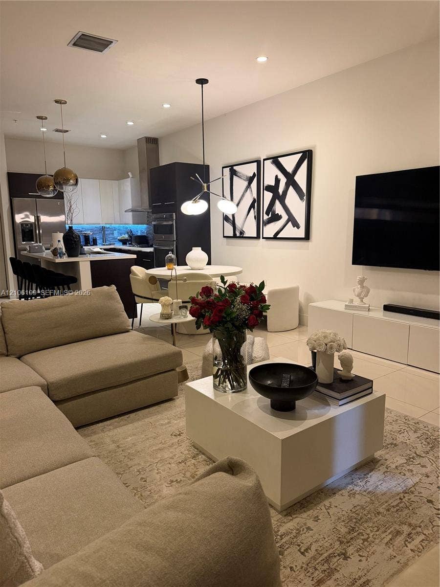 Open-concept kitchen featuring dark-toned cabinetry, stainless steel appliances, a center island with bar seating, pendant lighting, and neutral floor tiling