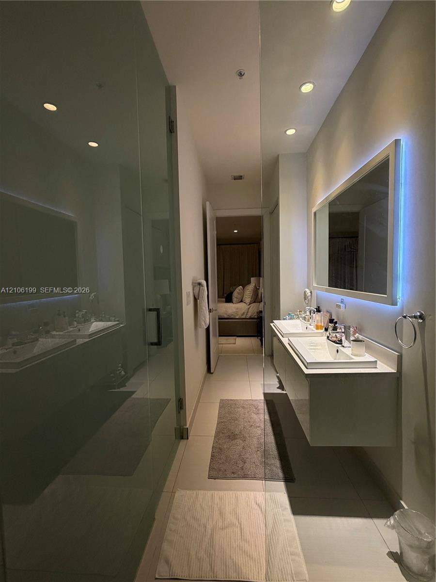 Floating dual-sink vanity with a rectangular mirror featuring integrated LED backlighting, glass-enclosed shower, recessed ceiling lighting, and large-format tile flooring