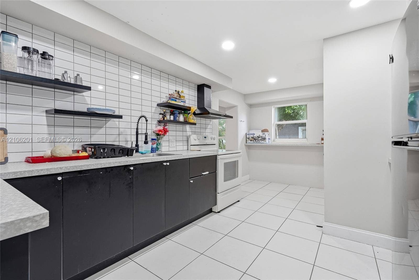 Kitchen featuring black flat-panel cabinetry, light-toned countertops, white subway tile backsplash, floating shelving, and white tile flooring