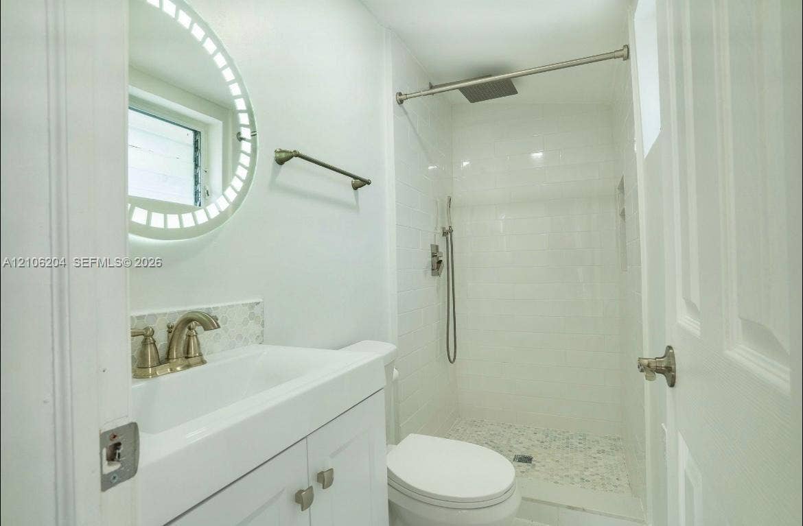 2121 SW 13th Avenue, #Studio, Miami, Miami, FL 33145 Bathroom featuring a white vanity cabinet, illuminated circular mirror, shower with white tiled surround, rain-style showerhead, and pebble-finish shower floor