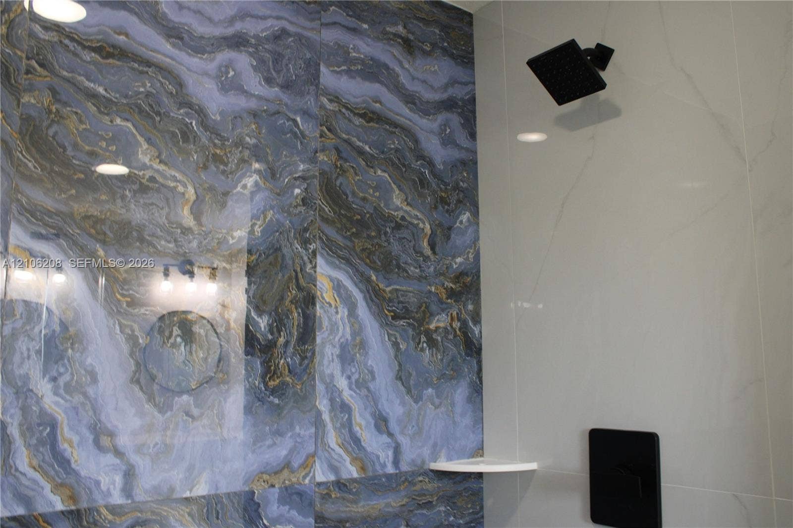 Large-format wall tile featuring a blue and gold marbled pattern, complemented by smooth white wall tile, matte black rain-style shower head, matching matte black pressure-balance valve, and an integrated corner shelf
