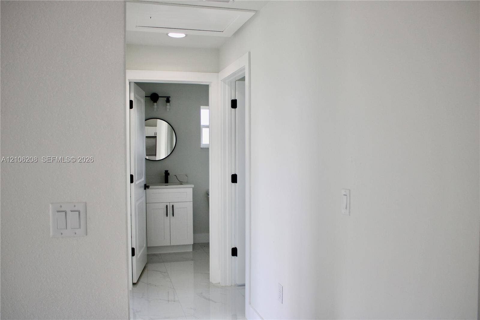 Hallway featuring light-colored tile flooring and recessed ceiling lighting, revealing a bathroom with a white vanity, matte black hardware, and a circular mirror