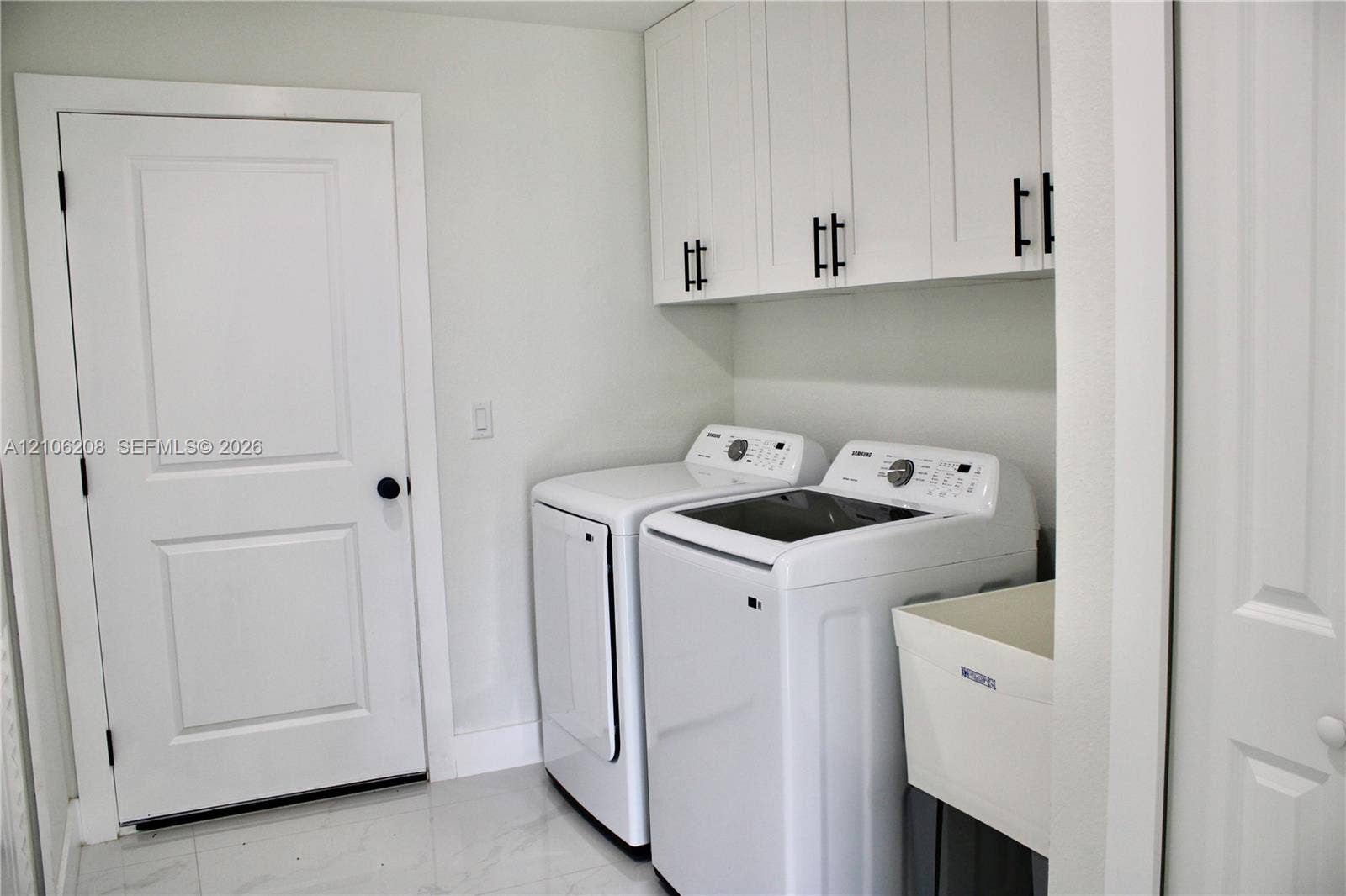 Laundry room featuring white shaker-style wall cabinets with matte black hardware, a utility sink, and large-format tile flooring