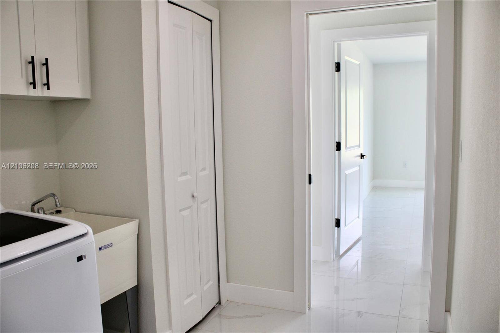 Utility space featuring white shaker-style wall cabinetry with matte black hardware, a basin sink, bifold door storage, and polished stone-finish floor tile