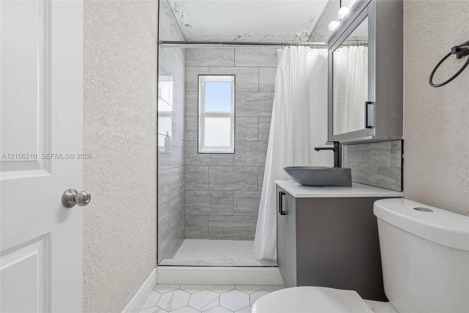 1111 S 7th Street, Fort Pierce, FL 34950 Bathroom featuring a shower stall with gray horizontal tile surround, a vanity with a dark cabinet and white countertop, a vessel sink with a matte black faucet, and white hexagonal floor tiling