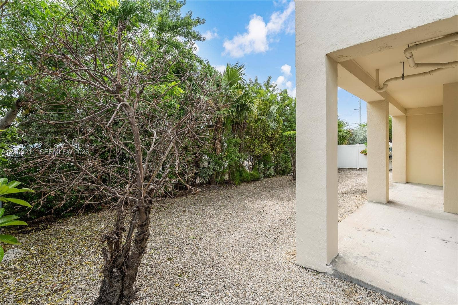 Covered concrete patio featuring structural support columns, gravel landscaping, and established perimeter vegetation