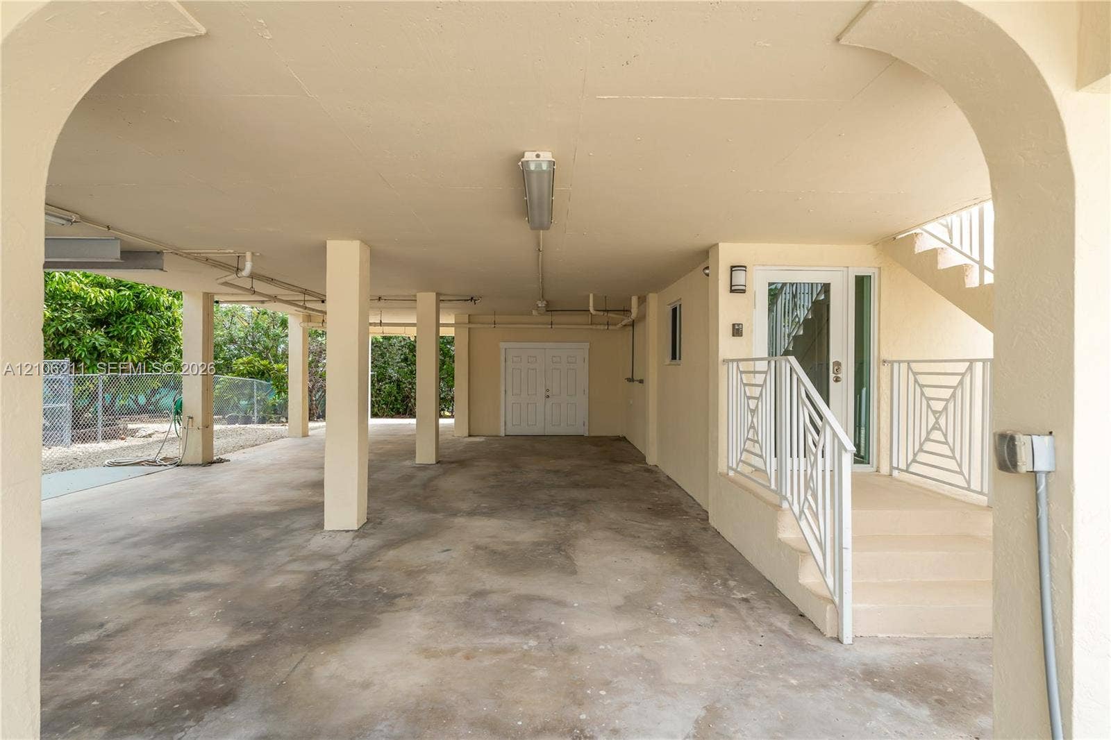 Covered carport with architectural archways, concrete flooring, dual-door utility entrance, and staircase with metal railings