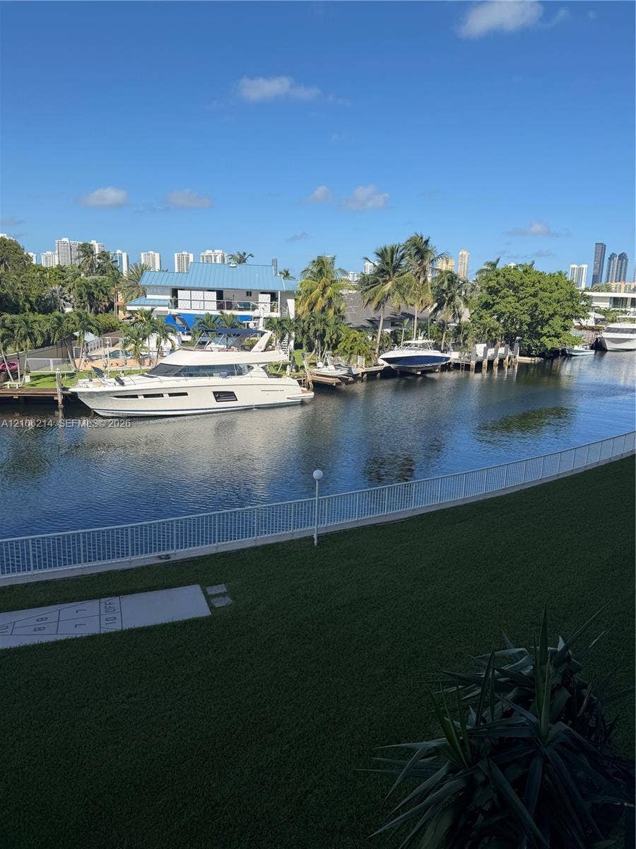2841 NE 163rd Street, #305, North Miami Beach, North Miami Beach, FL 33160 Waterfront property featuring a canal bulkhead, manicured lawn, white metal fencing, and a concrete hopscotch court
