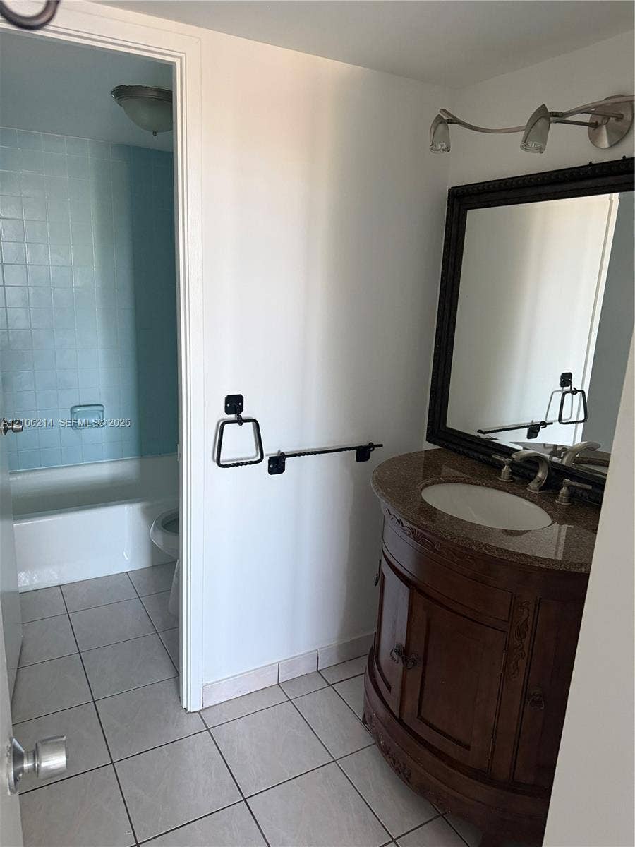 2841 NE 163rd Street, #305, North Miami Beach, North Miami Beach, FL 33160 Wood-finish vanity with a stone countertop, integrated undermount sink, multi-light fixture, framed wall mirror, and tile flooring