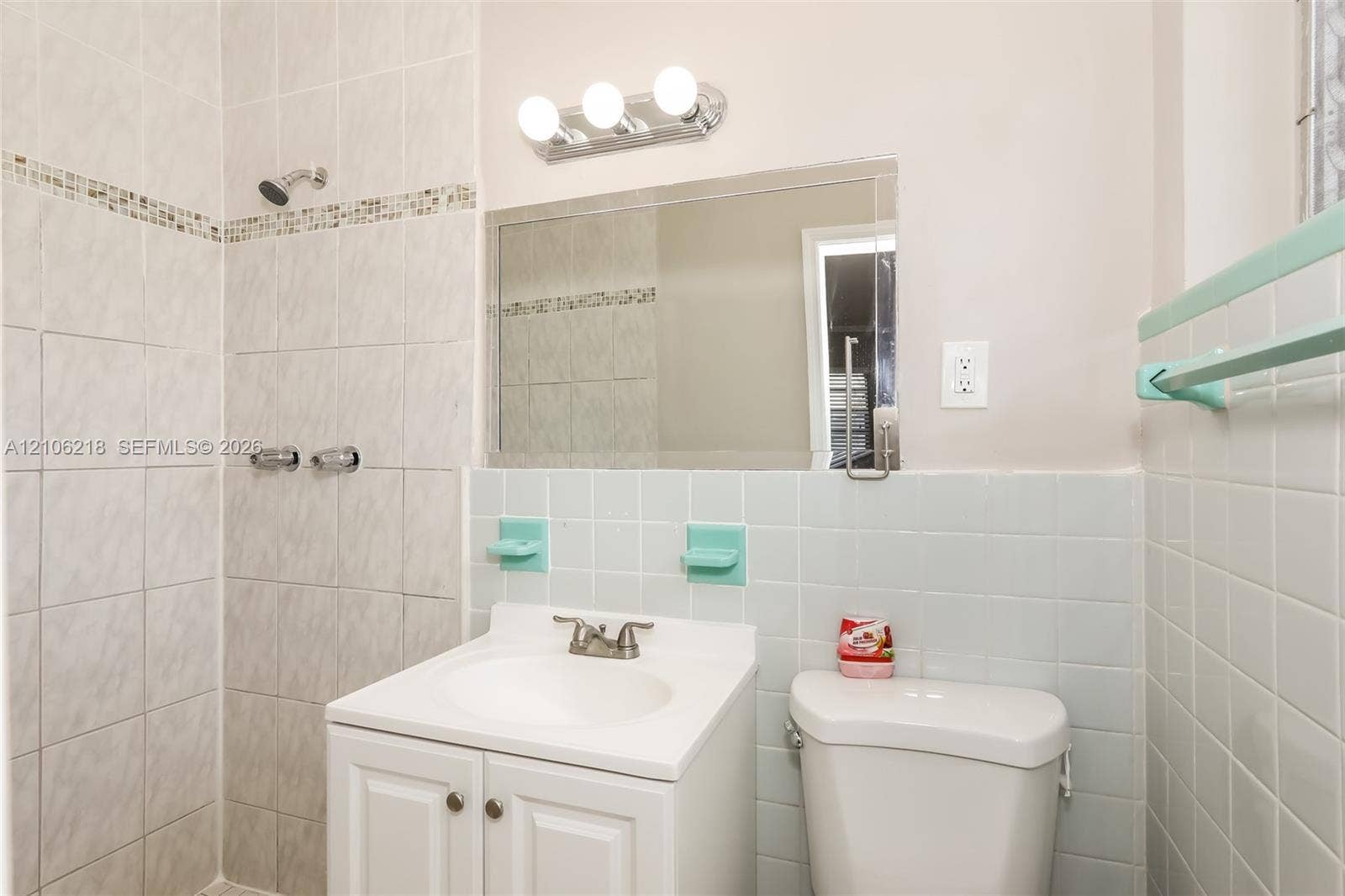 Bathroom featuring white cabinetry, a rectangular vanity sink with a metal faucet, wall-mounted three-light fixture, glass-paned mirror, and a tiled shower stall with a decorative accent border