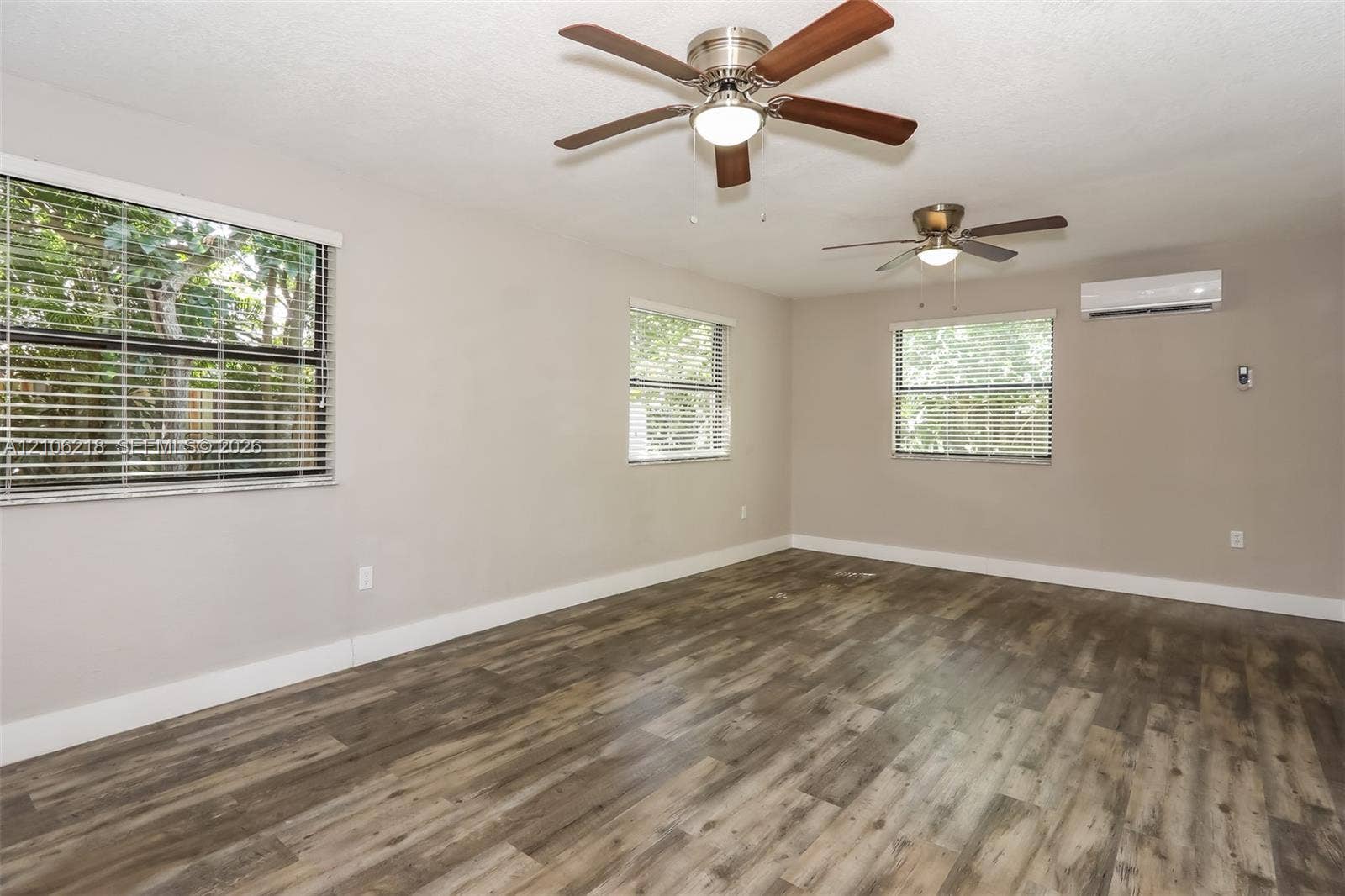 Spacious room featuring wood-finish flooring, two ceiling fans with integrated lighting, horizontal window blinds, and a wall-mounted ductless climate control unit