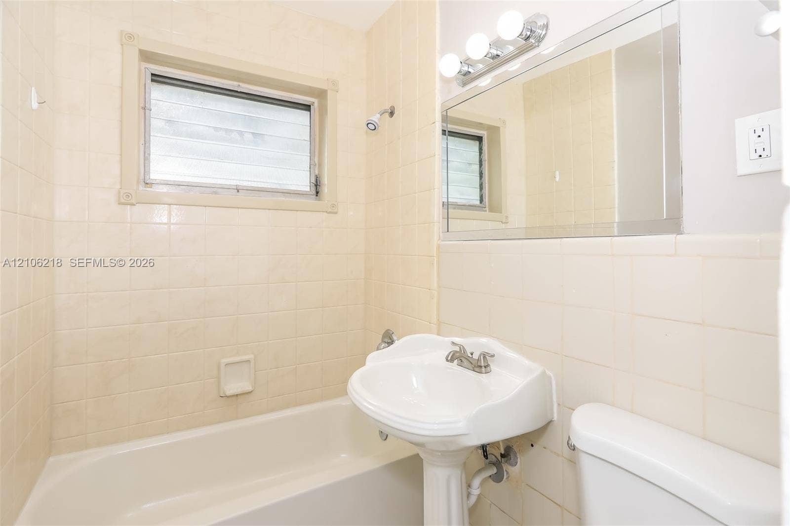 Bathroom featuring ceramic wall tiling, pedestal sink, acrylic soaking tub, built-in recessed soap dish, and vanity mirror with integrated lighting