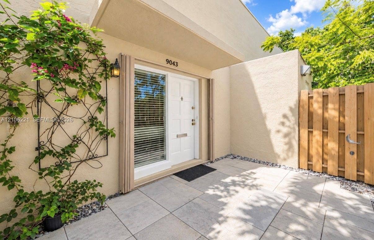Entryway featuring a paved courtyard, stucco exterior, decorative metal trellis, wood-panel privacy fencing, and a white paneled front door