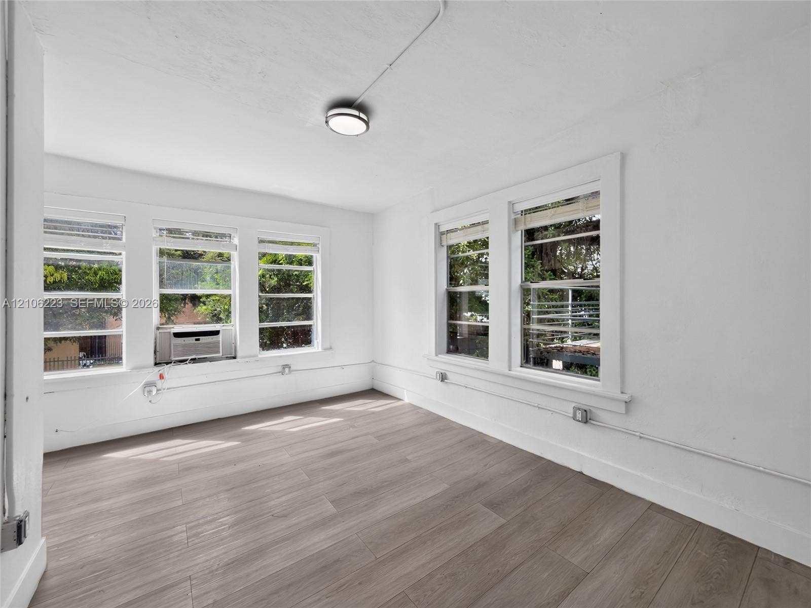 Bright corner room featuring expansive multi-pane windows flooding the space with natural light, wood-finish flooring, clean white walls, and recessed ceiling lighting