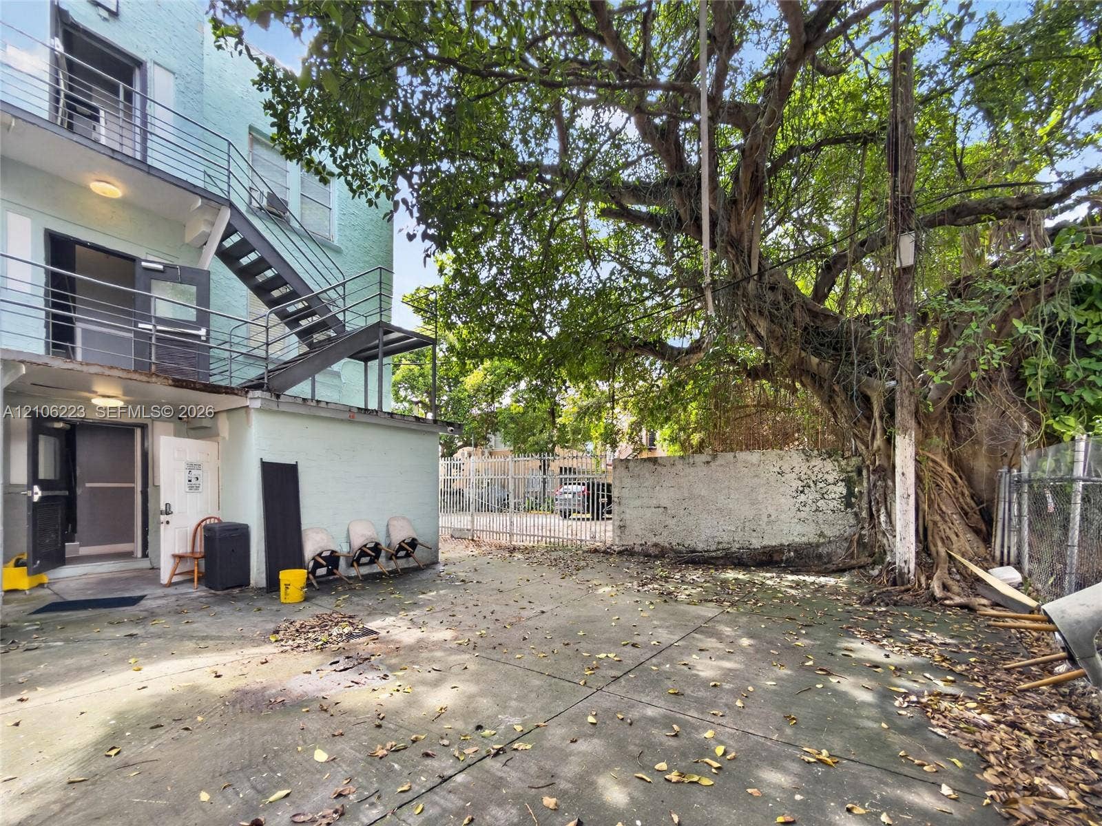 Concrete exterior courtyard featuring a multi-level metal fire escape, masonry wall, mature tree, and chain-link perimeter fencing