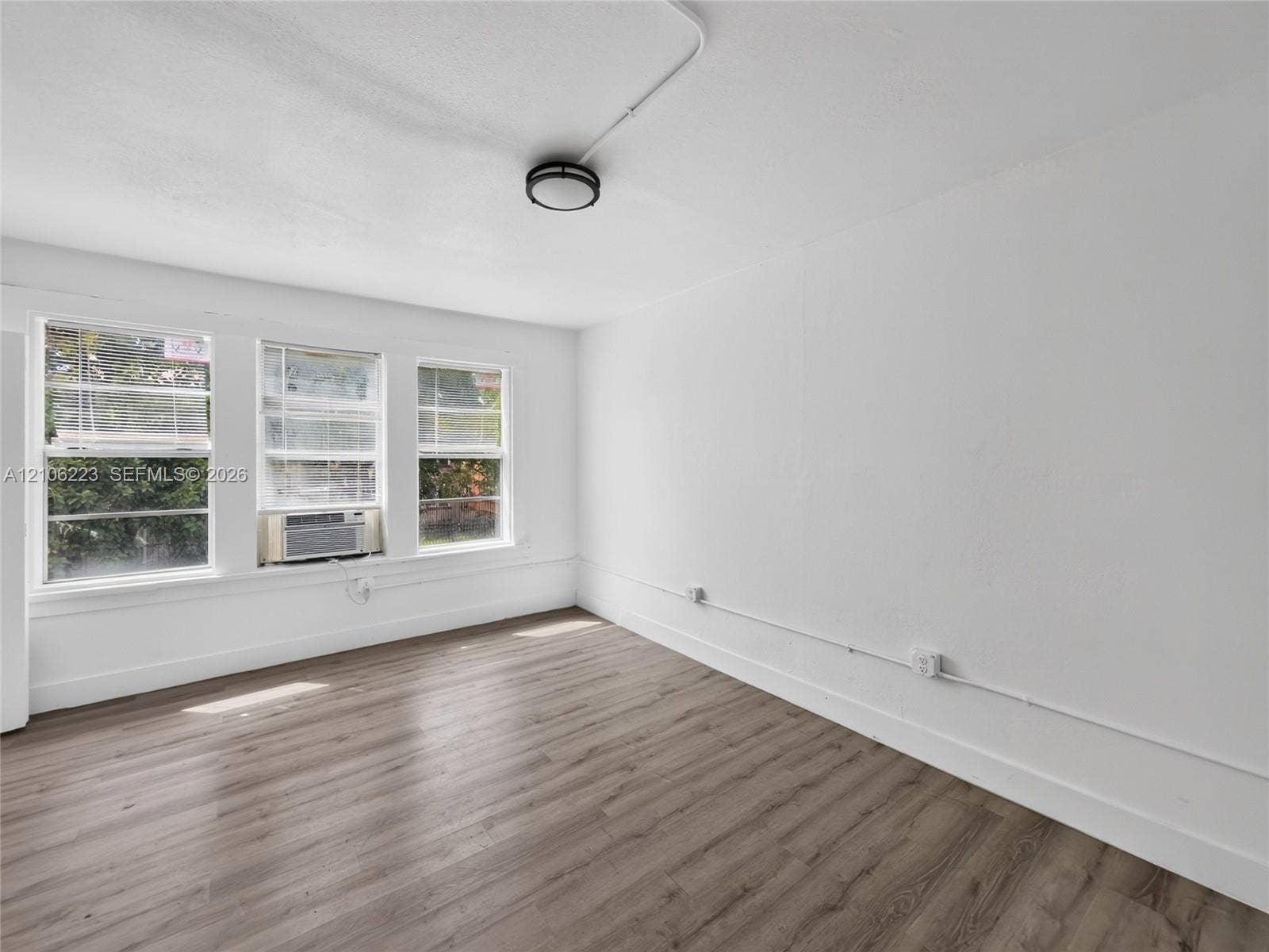 Interior space featuring wood-finish flooring, white walls, triple window configuration, and flush-mount ceiling lighting