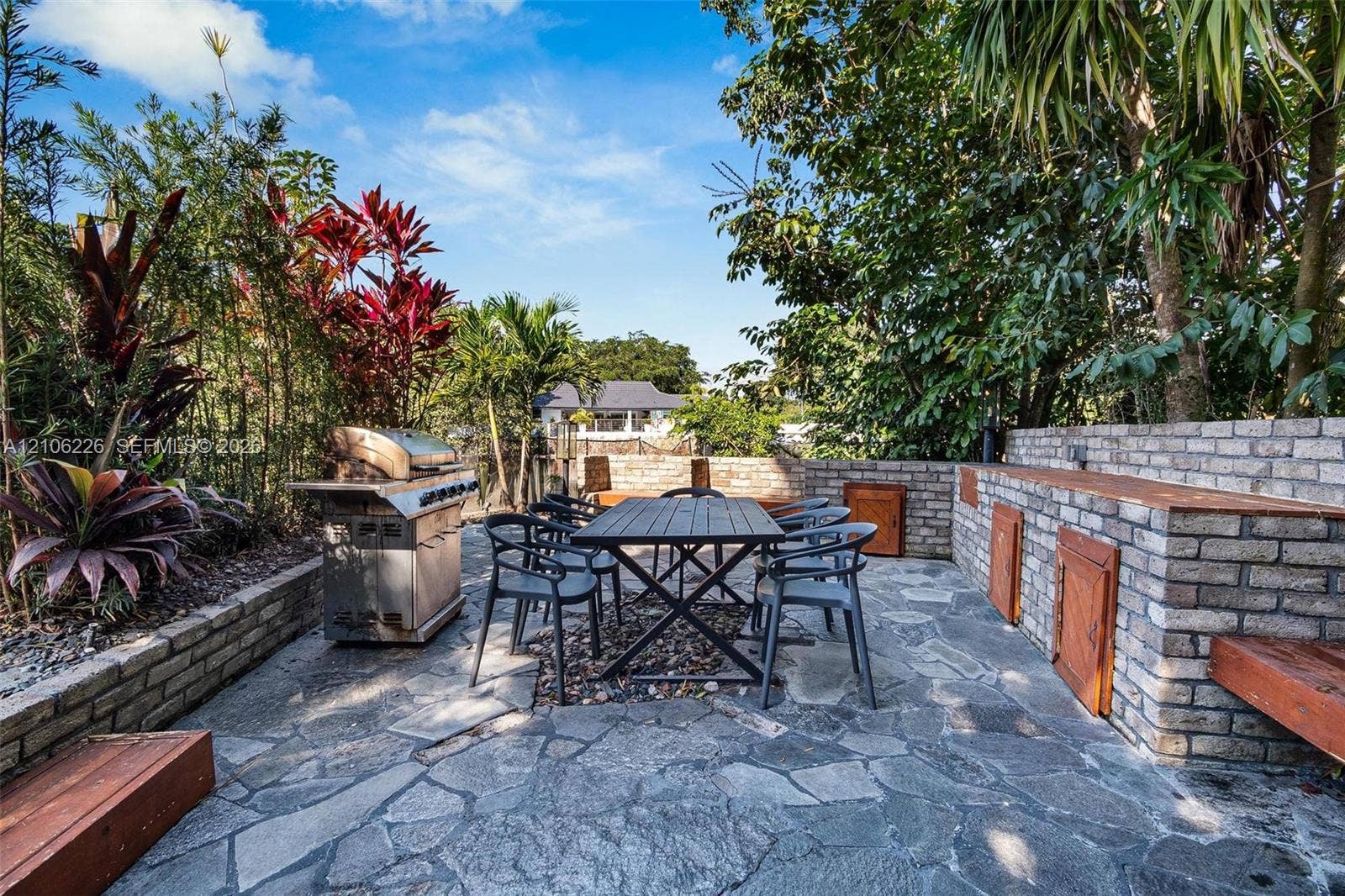 11155 SW 40th Street, Davie, FL 33328 Outdoor patio featuring irregular flagstone paving, a built-in brick grilling station, integrated wood-bench seating, and established tropical landscaping