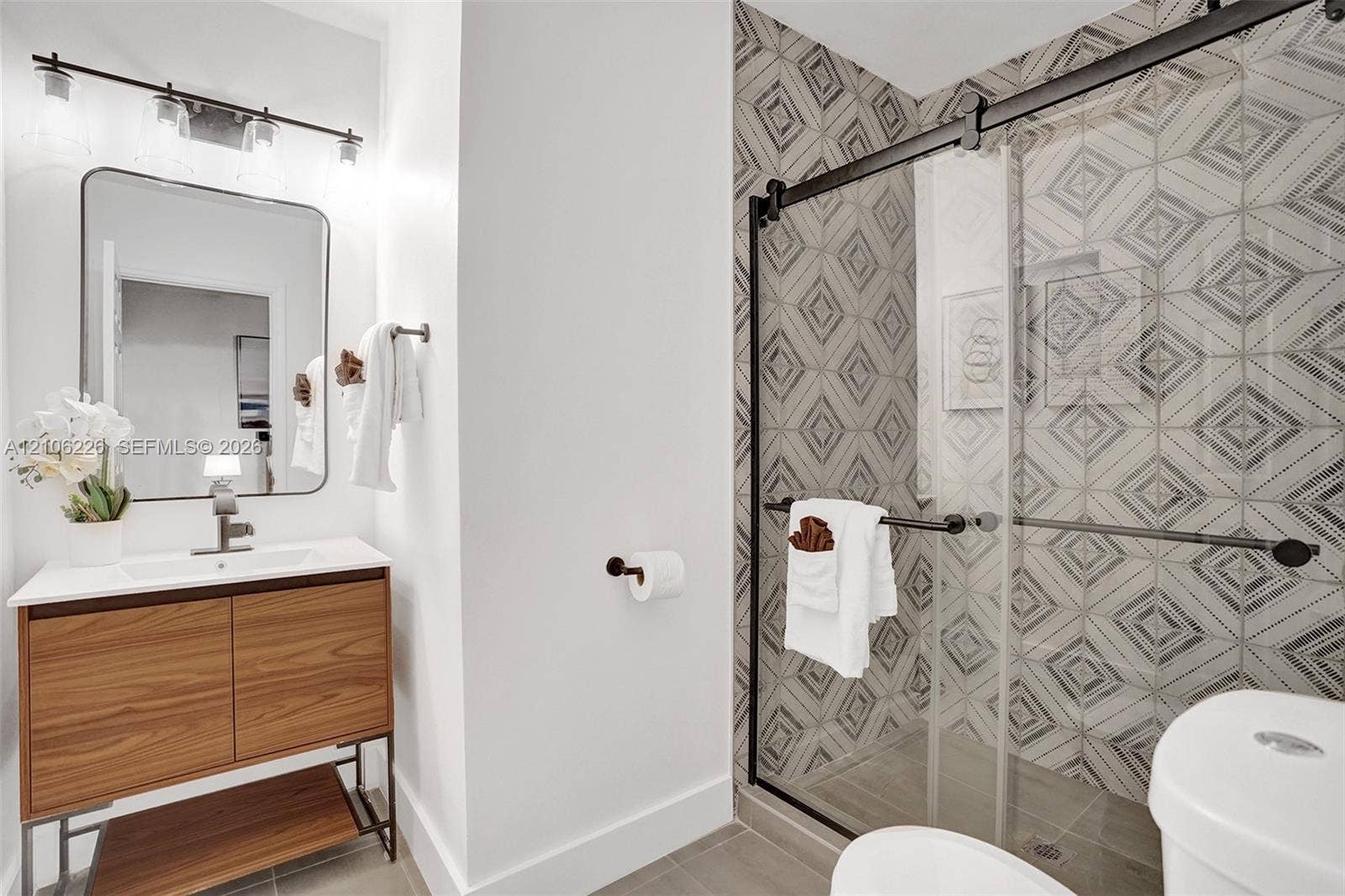 11155 SW 40th Street, Davie, FL 33328 Bathroom featuring a wood-finish floating vanity with a white countertop, an integrated rectangular sink, a glass-enclosed shower with geometric patterned tile walls, and matte black hardware