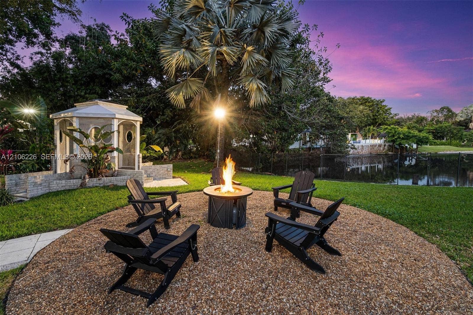 11155 SW 40th Street, Davie, FL 33328 Landscaped backyard featuring a circular gravel fire pit area, stone-surround gazebo, manicured lawn, and water frontage