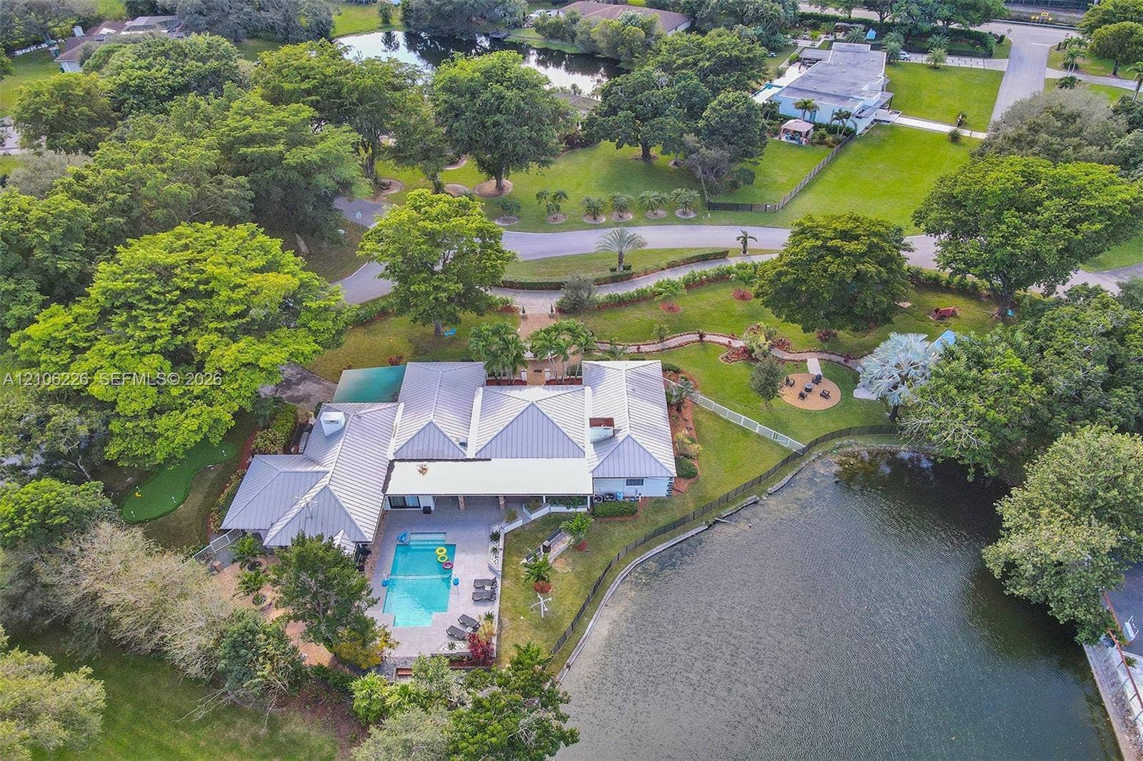 11155 SW 40th Street, Davie, FL 33328 Aerial perspective featuring a metal roof structure, in-ground swimming pool with stone-finish decking, and extensive waterfront grounds with mature canopy trees