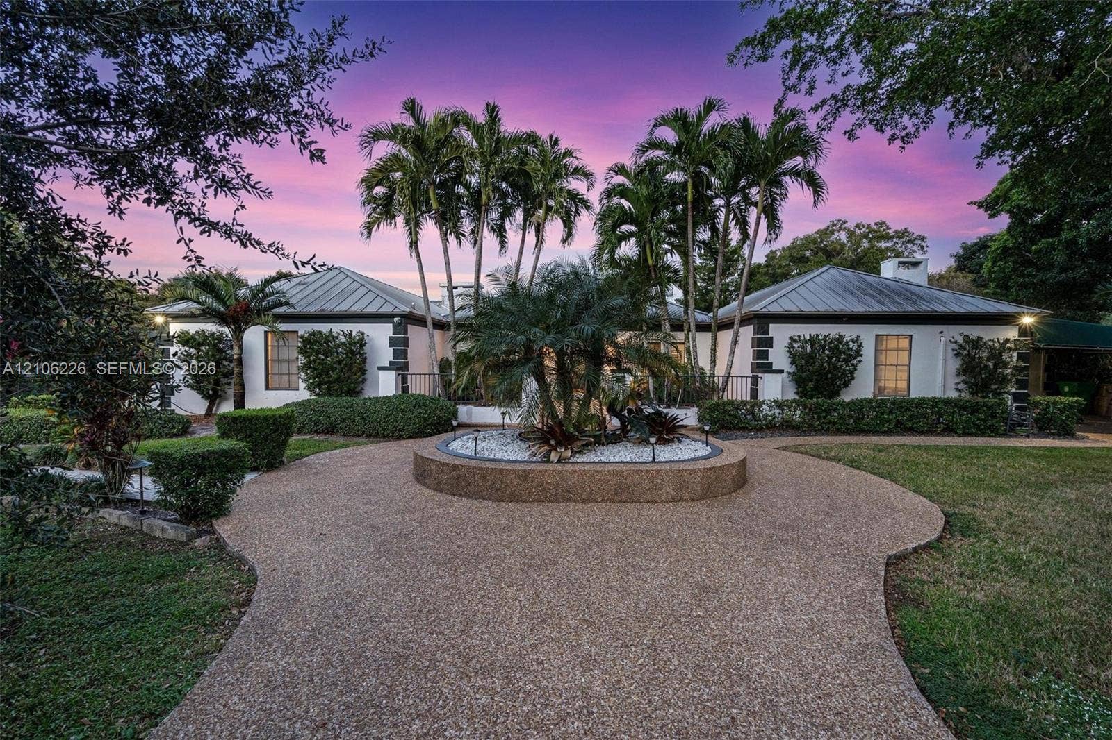 11155 SW 40th Street, Davie, FL 33328 Single-story residence featuring a metal roof, aggregate stone driveway, central circular garden bed with white stone mulch, and mature palm landscaping