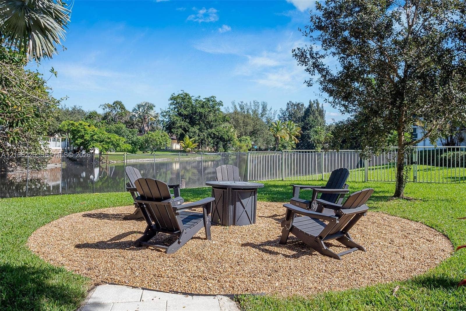 11155 SW 40th Street, Davie, FL 33328 Landscaped backyard featuring a circular gravel fire pit area, manicured lawn, metal perimeter fencing, and mature trees overlooking a pond