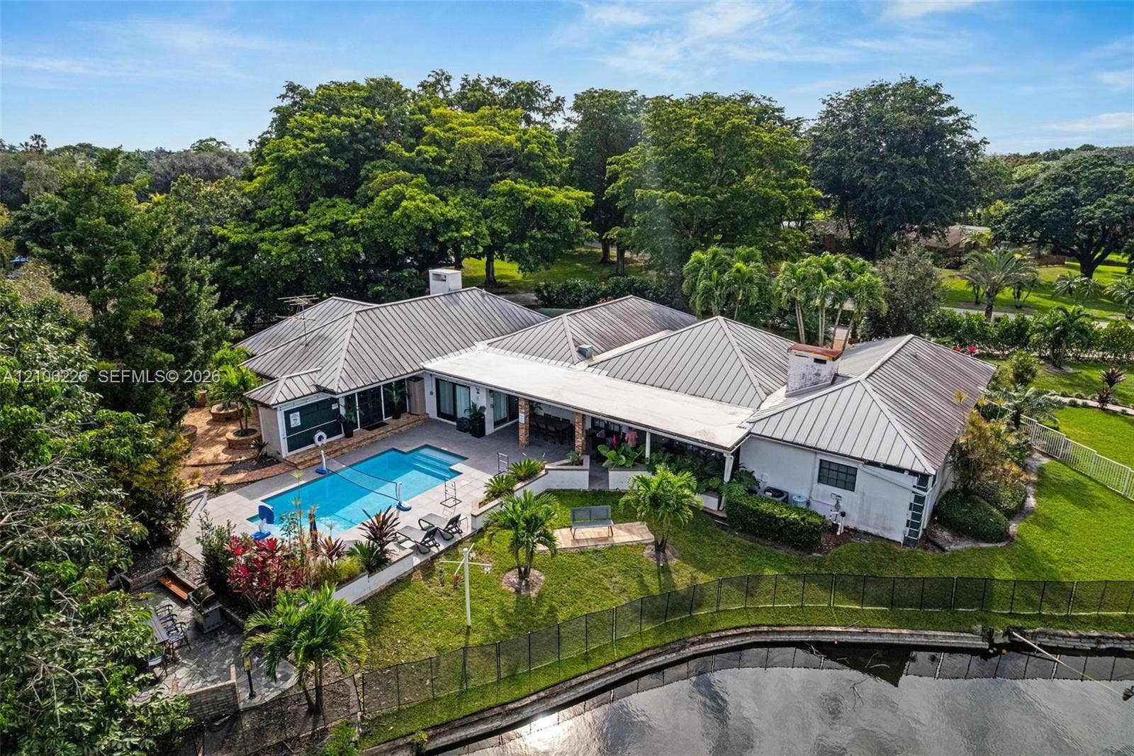 11155 SW 40th Street, Davie, FL 33328 Metal roof residence featuring an in-ground swimming pool, covered patio, waterfront grounds, and mature landscaping