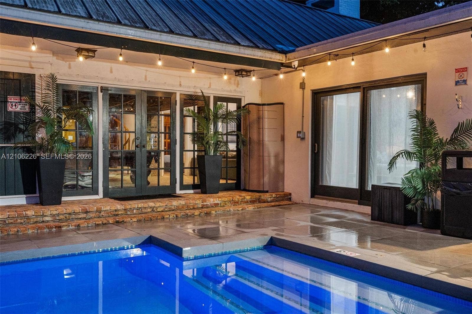 11155 SW 40th Street, Davie, FL 33328 Outdoor pool area featuring a stone patio, brick-bordered raised platform, multi-panel glass entry doors, and sliding glass doors
