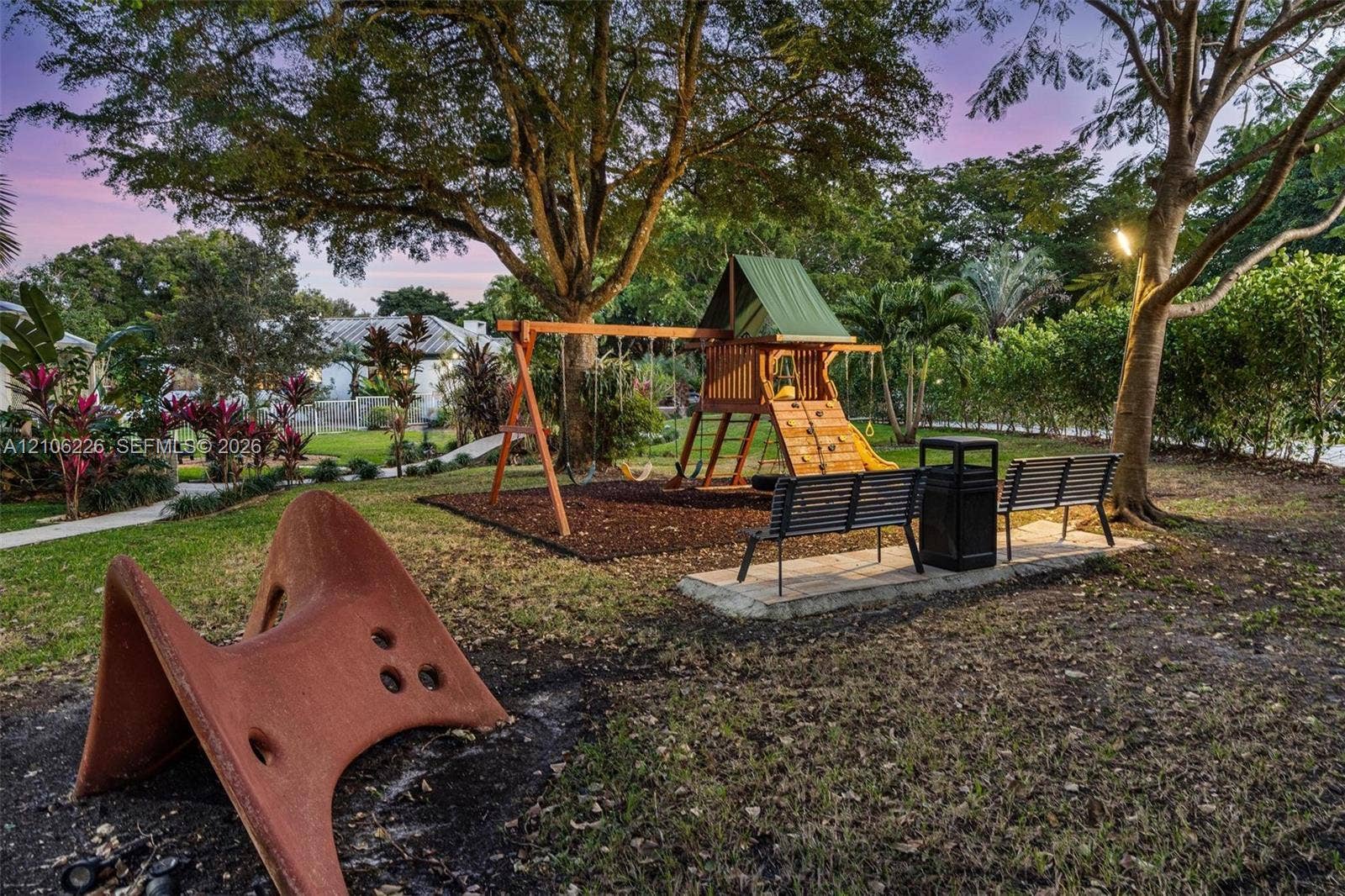 11155 SW 40th Street, Davie, FL 33328 Expansive outdoor grounds featuring a wooden play structure, mature trees, manicured landscaping, paved walkways, and metal bench seating