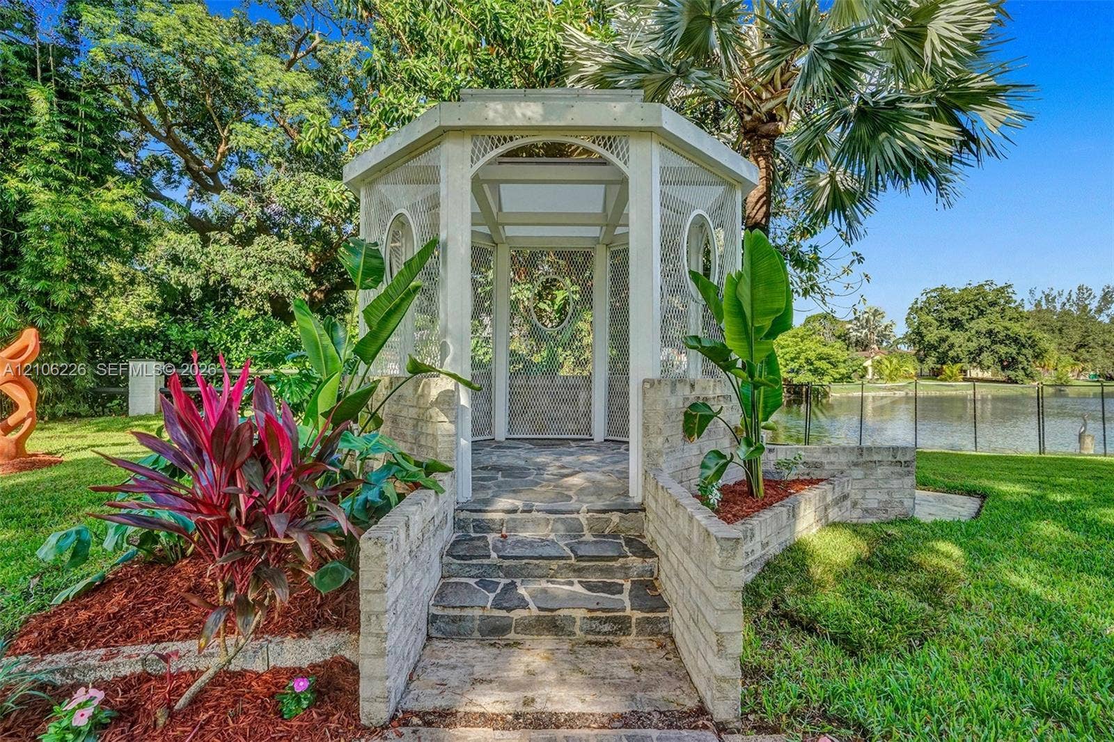 11155 SW 40th Street, Davie, FL 33328 Octagonal screened gazebo featuring a stone-clad foundation, masonry steps, white-painted framework, and landscaped garden beds