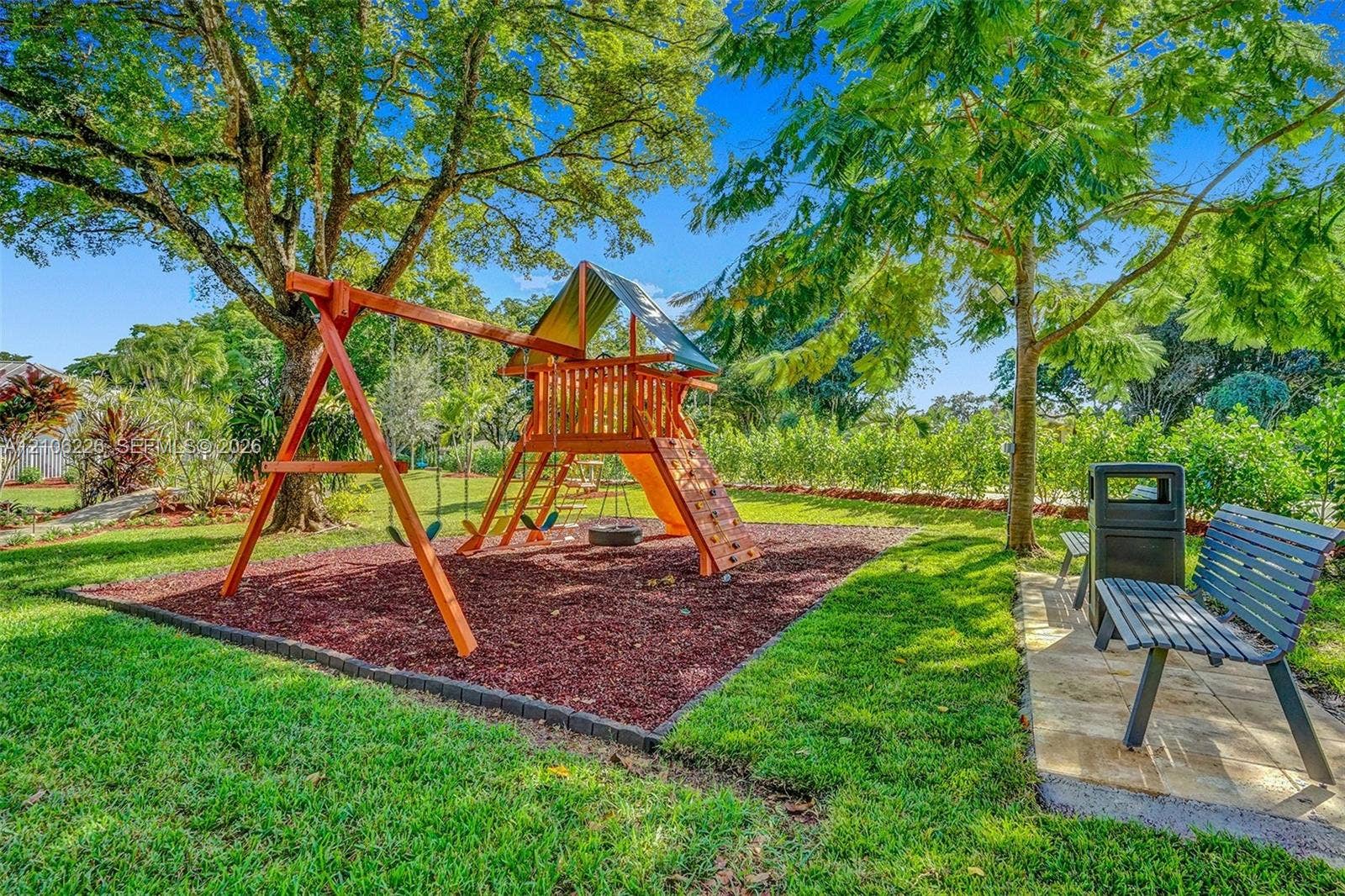 11155 SW 40th Street, Davie, FL 33328 Outdoor recreational space featuring a wooden play structure, a mulched activity area, a stone-paved walkway, and mature shade trees