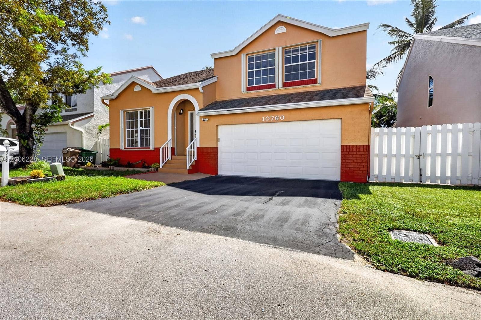 10760 SW 11th Drive, Davie, FL 33324 Two-story exterior featuring a stucco facade, an arched entryway with metal railing, a two-car garage, and a white privacy fence
