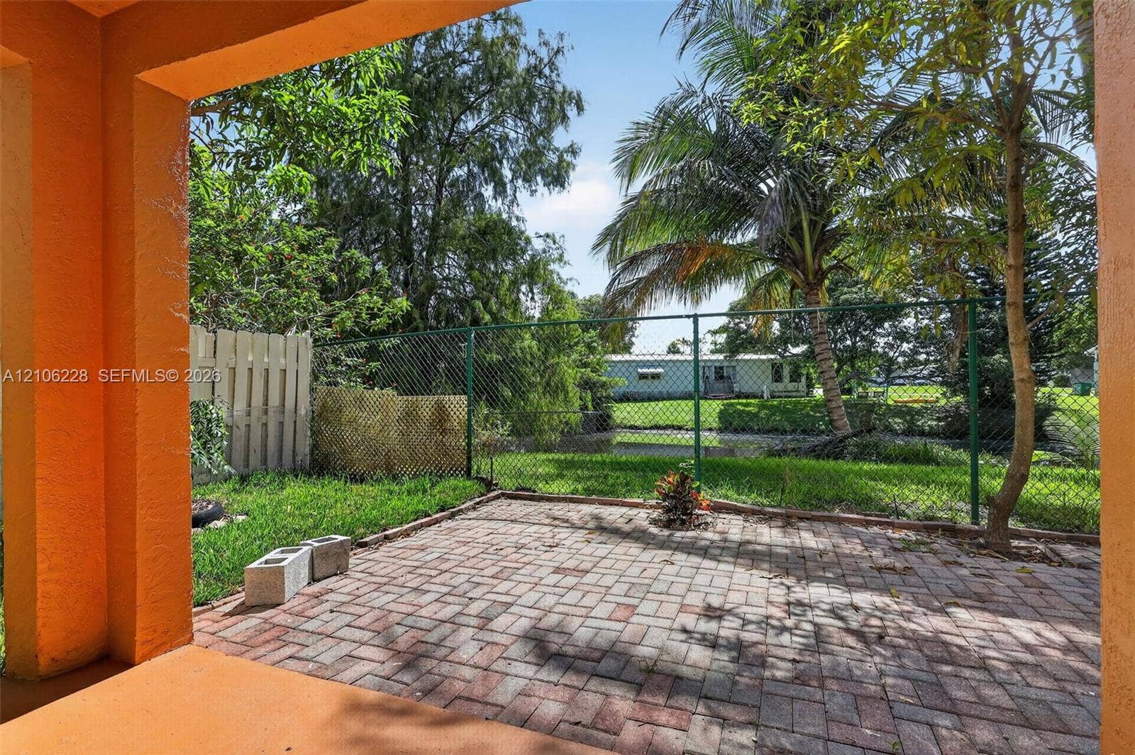 10760 SW 11th Drive, Davie, FL 33324 Covered patio featuring brick pavers, chain-link fencing, manicured lawn, and mature palm trees