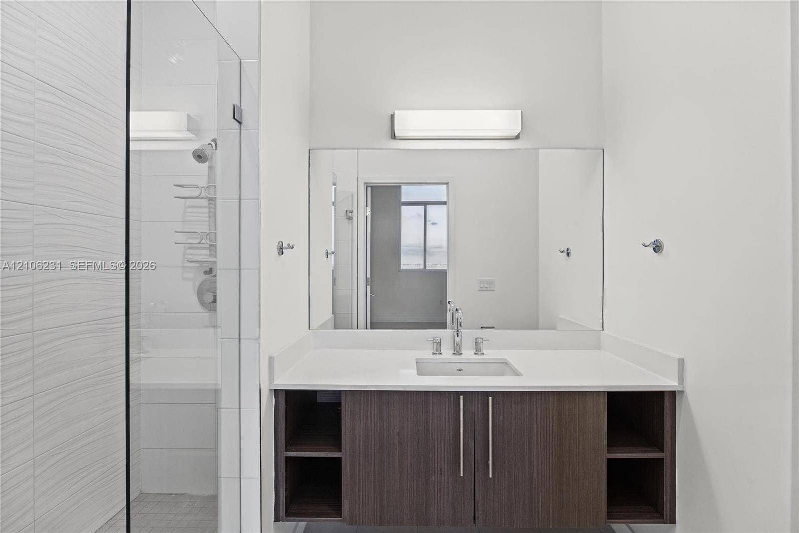 7875 NW 107th Avenue, #802, Doral, FL 33178 Private bathroom with wall-mounted vanity, wood-finish cabinetry with open side shelving, white countertop with an integrated rectangular sink, a polished chrome faucet, a large frameless mirror, and a glass-enclosed shower with textured wall tile