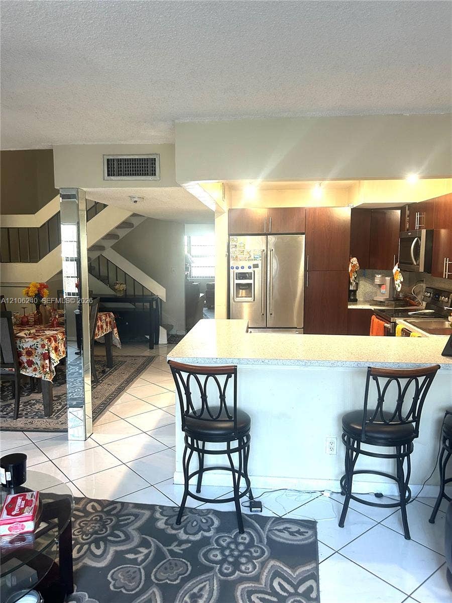 2554 N 38th Avenue, Hollywood, FL 33021 Kitchen featuring wood cabinetry, stainless steel appliances, and a breakfast bar with light-toned stone-finish countertop, accented by polished mirrored support pillars and white tile flooring