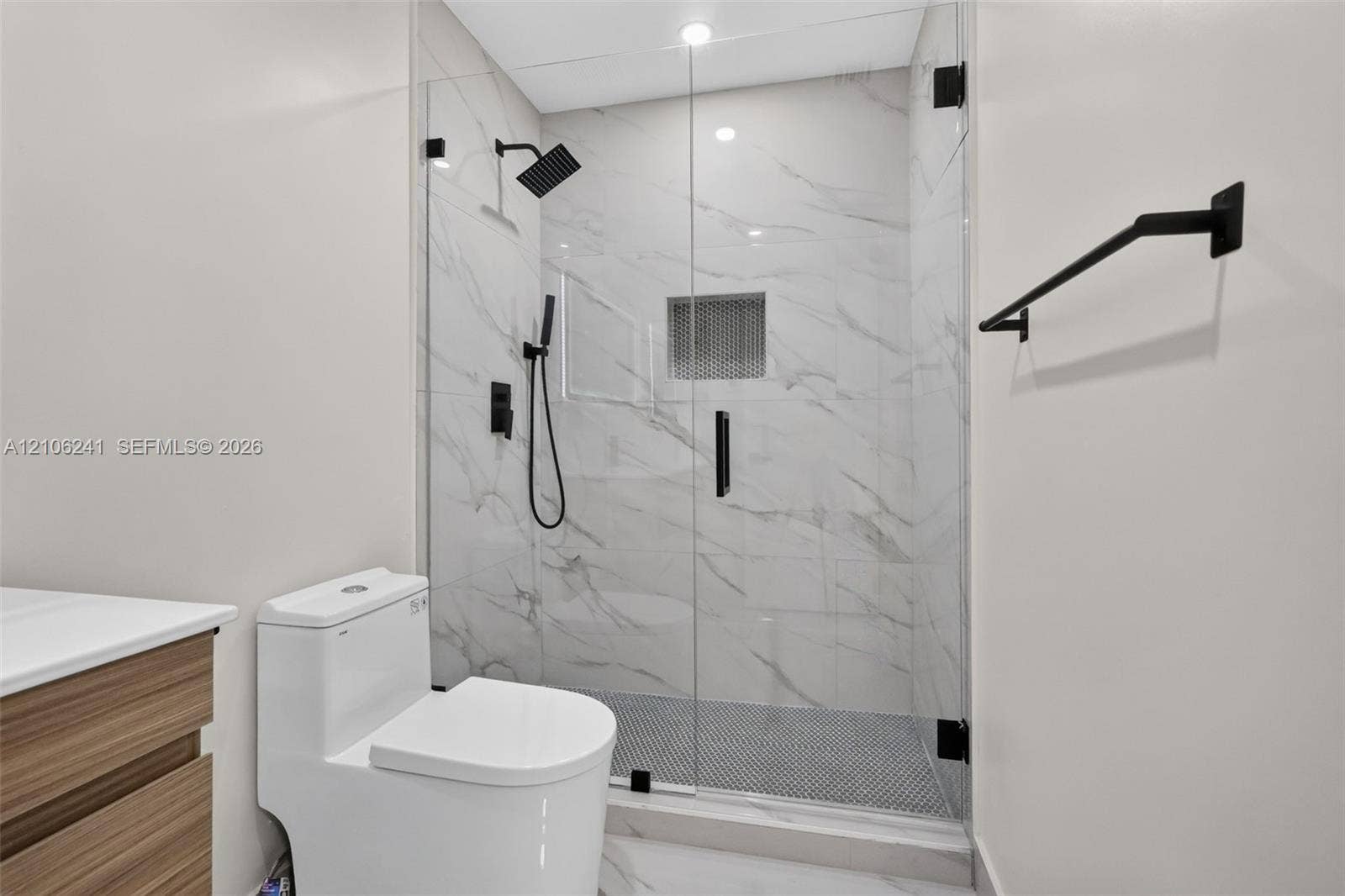 9407 SW 18th Street, Miramar, FL 33025 Walk-in shower featuring a frameless glass enclosure, floor-to-ceiling stone-patterned wall tile, matte black rainfall showerhead with handheld sprayer, mosaic-tiled shower floor, and a built-in recessed wall niche