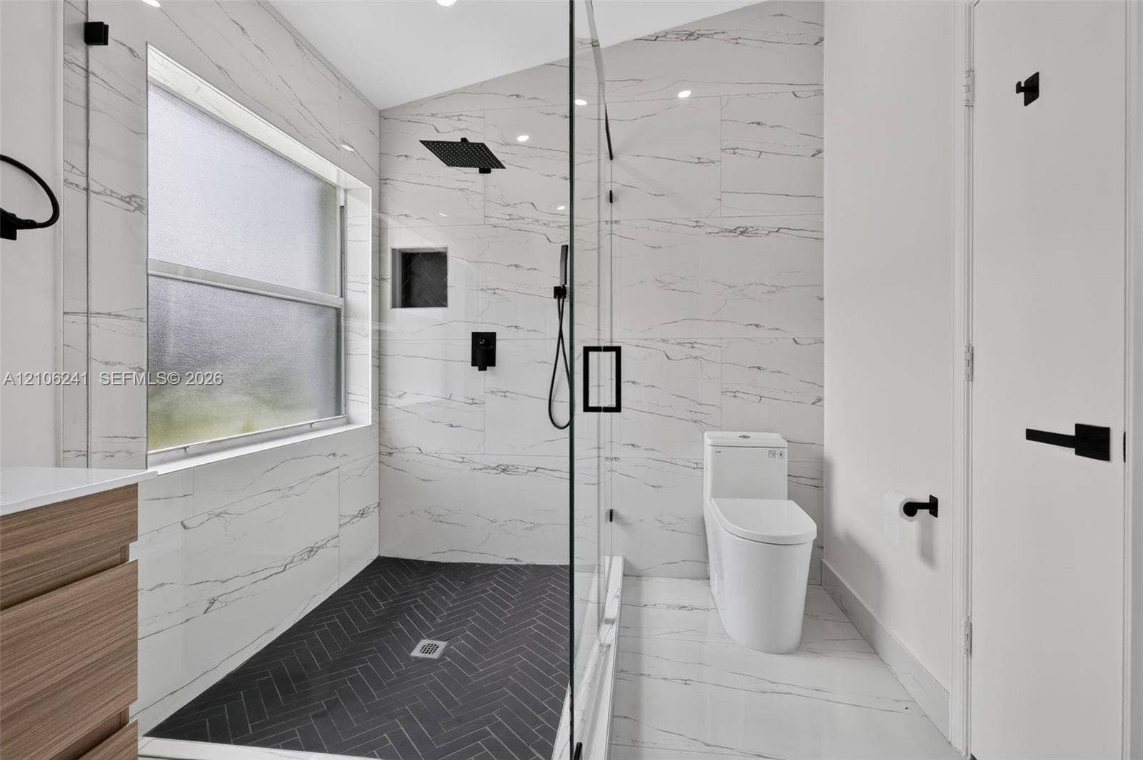 9407 SW 18th Street, Miramar, FL 33025 Walk-in glass-enclosed shower featuring dark herringbone floor tile, white veined wall tile, built-in recessed shelf, and matte black rainfall showerhead