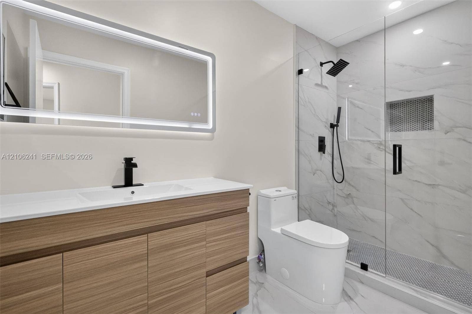 9407 SW 18th Street, Miramar, FL 33025 Wood-finish floating vanity with a stone countertop, backlit rectangular mirror, walk-in shower with stone-surround walls, matte black fixtures, and a glass shower door