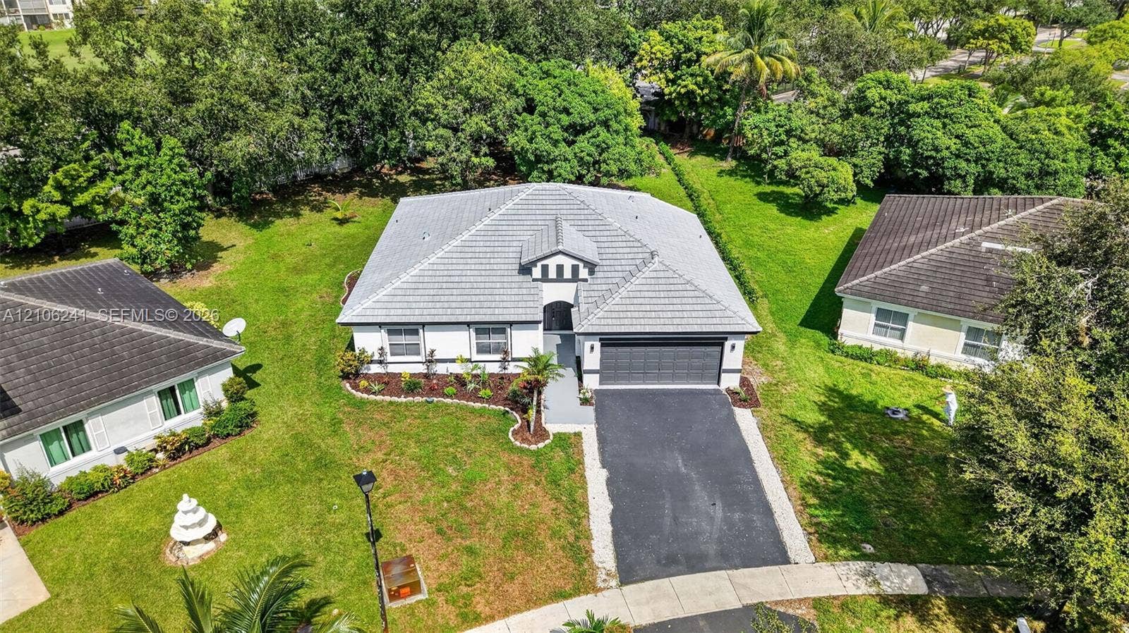 9407 SW 18th Street, Miramar, FL 33025 Single-story residence featuring a tile roof, light-toned exterior finish, attached two-car garage, asphalt driveway, and surrounding lawn with mature landscaping