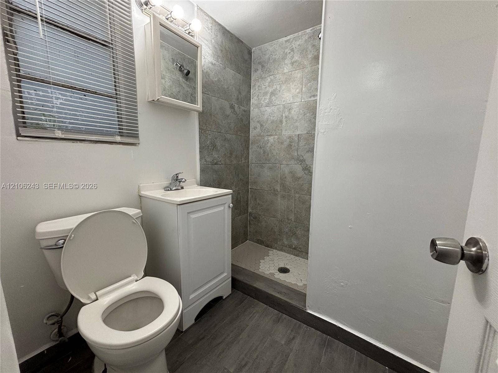 Bathroom featuring a walk-in shower with grey stone-finish tile surround, wood-finish flooring, white vanity cabinet, and mirrored medicine cabinet