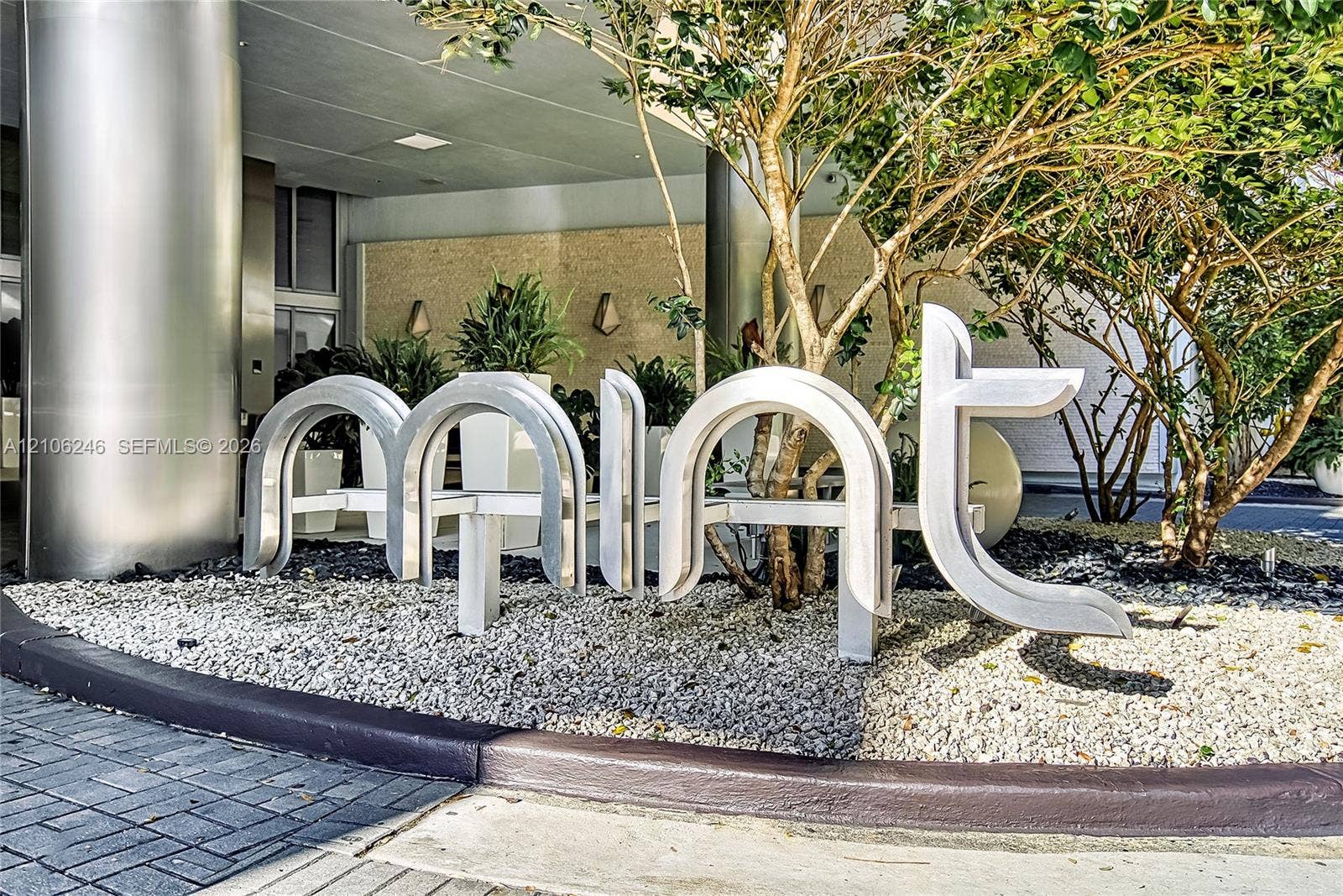 92 SW 3rd Street, #1511, Miami, Miami, FL 33130 Exterior entrance featuring brushed metal structural columns, stone-textured facade wall, sculptural metal signage, and gravel landscaping with integrated greenery