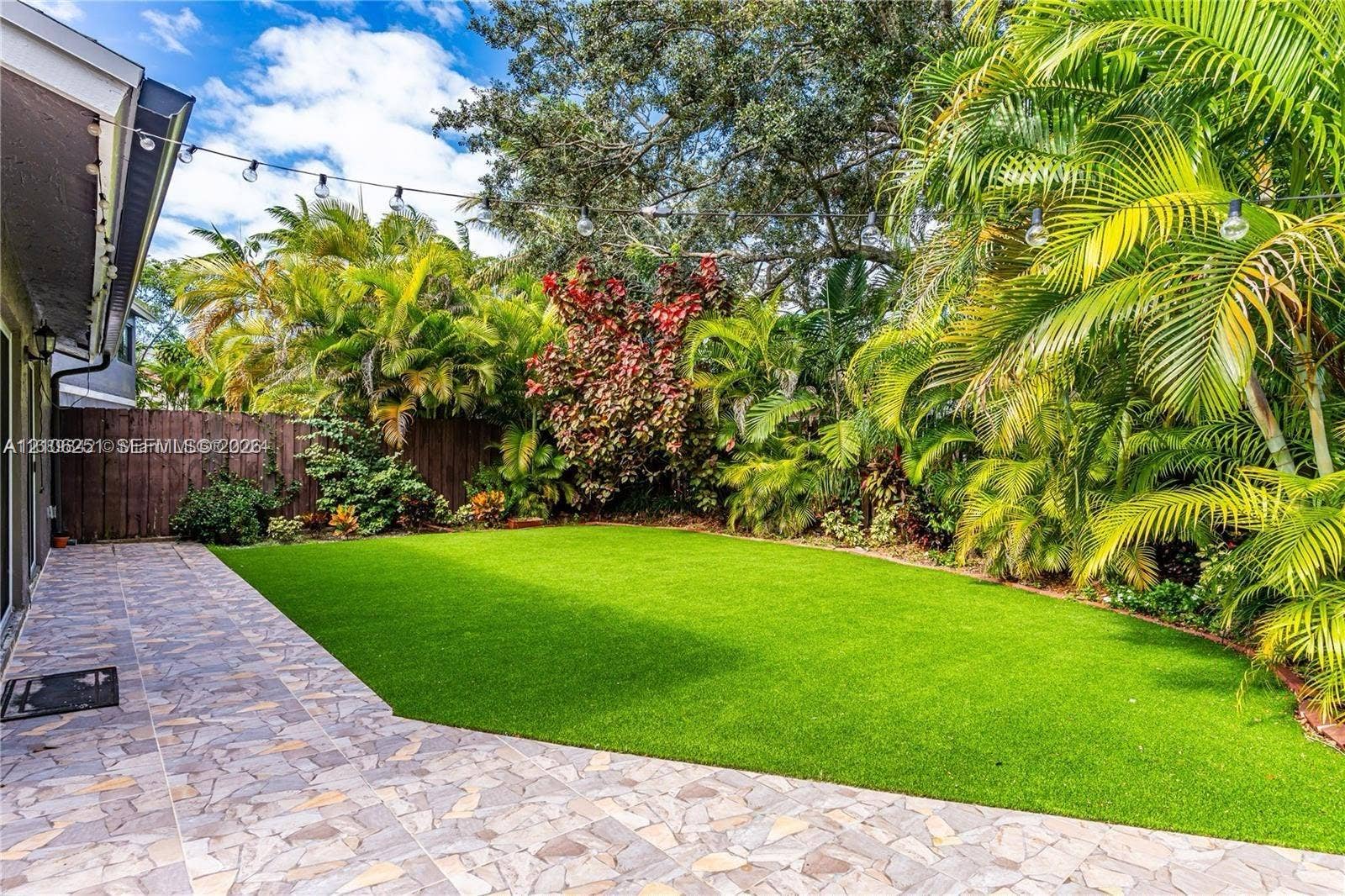 Backyard featuring a paved stone patio, synthetic turf lawn, wood privacy fencing, and established tropical landscaping