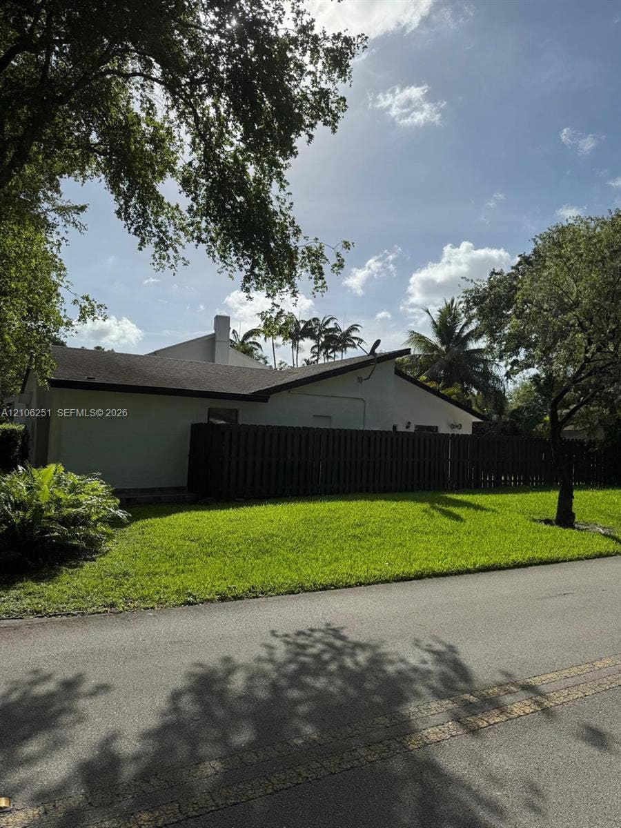 Stucco exterior residence featuring a dark wood-tone privacy fence, paved asphalt street access, and established mature trees