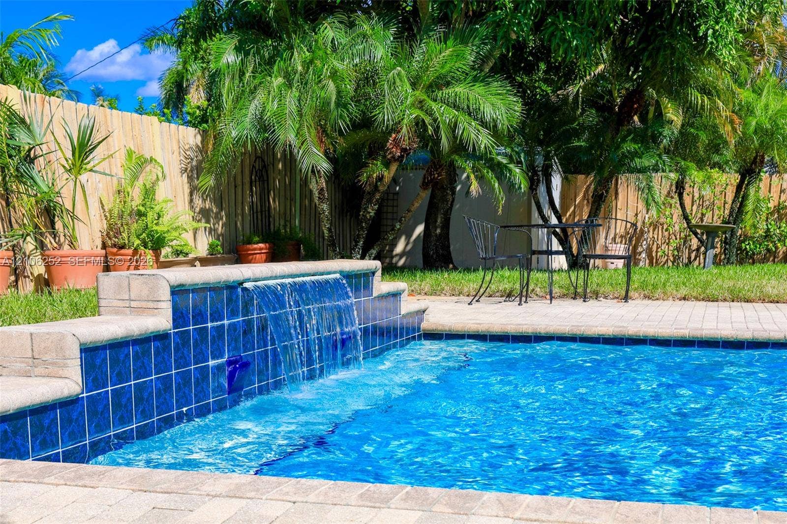 225 E Ilex Drive, Lake Park, Lake Park, FL 33403 Swimming pool featuring a tiled waterfall wall, paver decking, and surrounding mature palm trees within a fenced perimeter