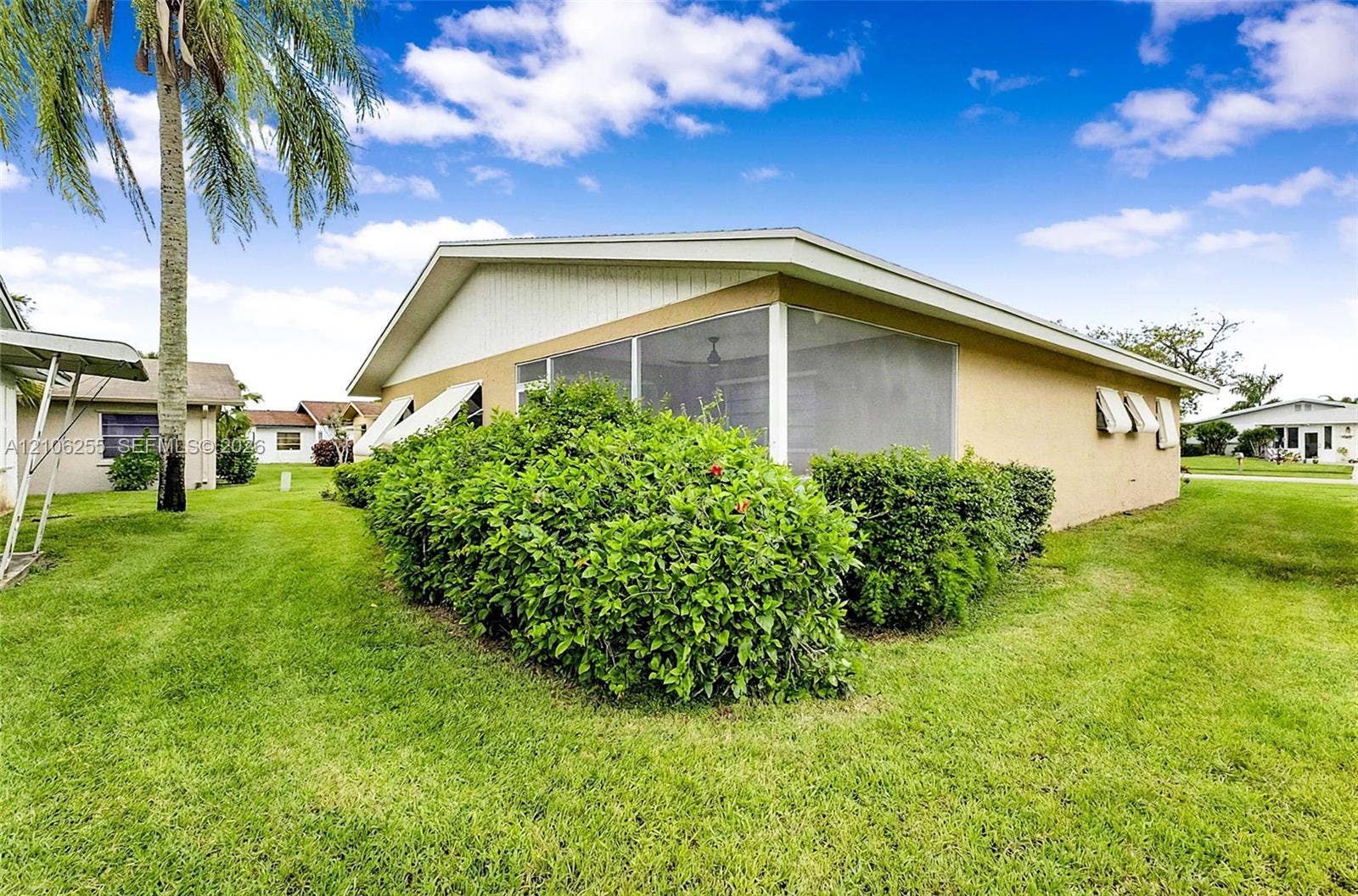 6118 Dusenburg Road, Delray Beach, FL 33484 Stucco exterior residence featuring a screened-in lanai, decorative window awnings, and landscaped perimeter shrubbery set on a level lawn